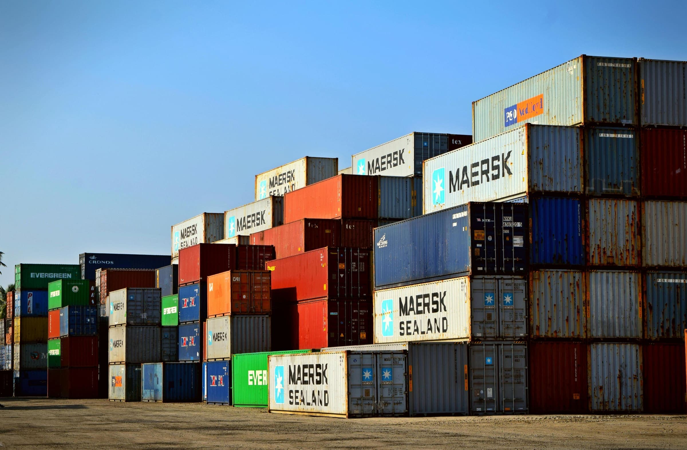 Rows of shipping containers stacked at a port under a blue sky